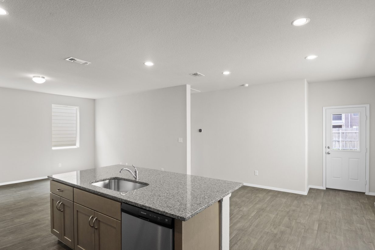 Interior image of kitchen with tan cabinets and granite countertops and living room with vinyl flooring.
