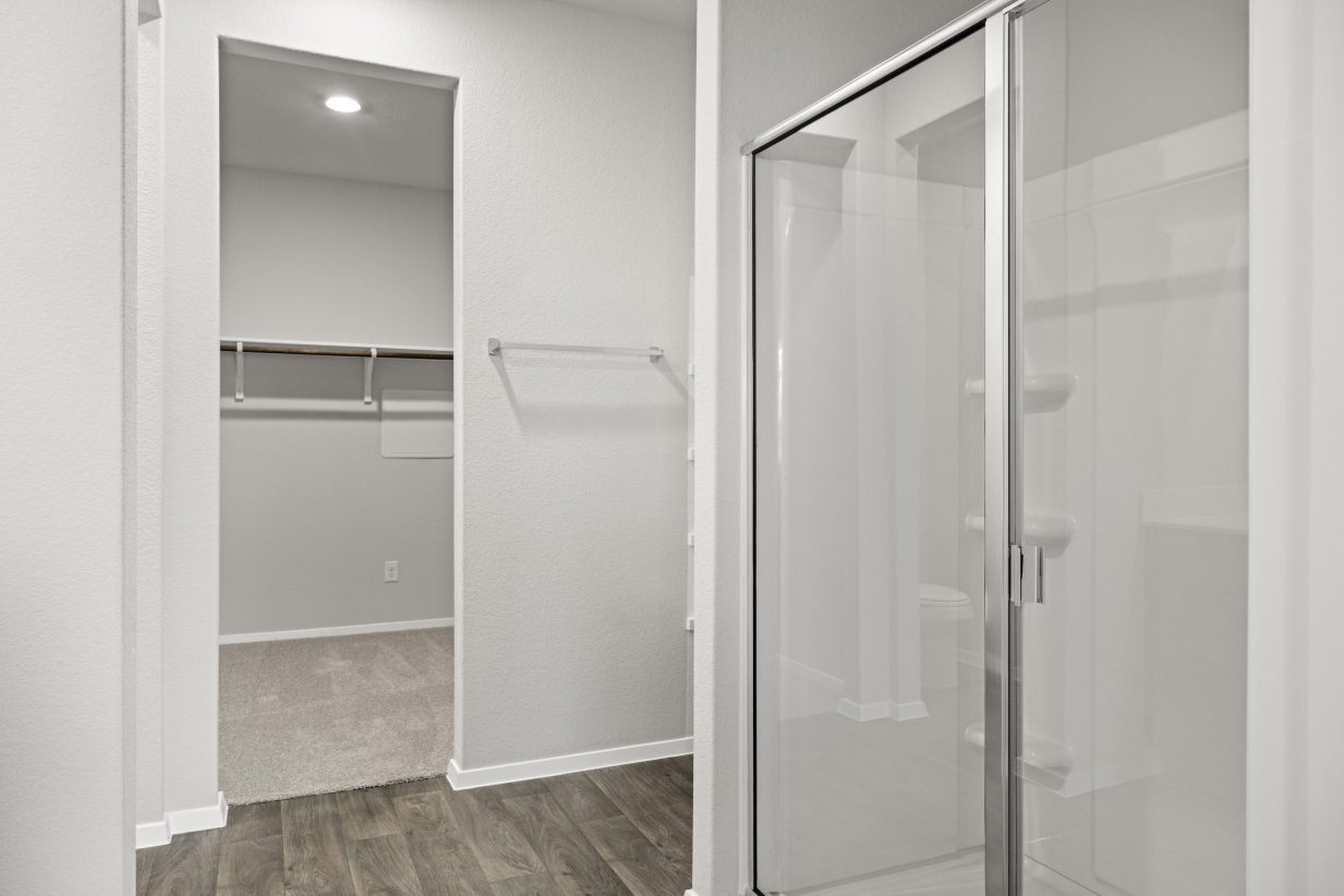A bathroom with a glass walk-in shower, towel rack, vinyl flooring, and white painted walls looking into the walk-in closet with brown carpet.