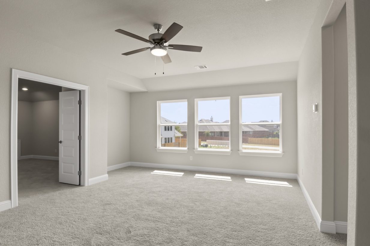The primary bedroom with three white-framed windows, tan carpet, a brown ceiling fan and a open door.