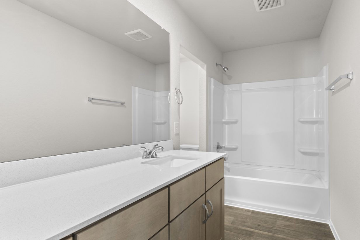 Interior image of a bathroom with a vanity with white countertops and brown cabinets, white walls, a mirror, and a white tub.