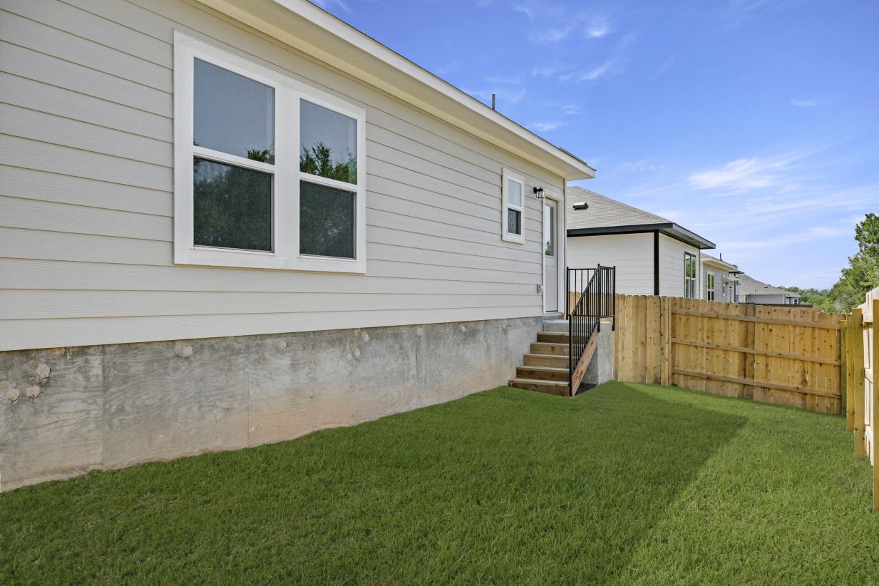 Backyard of gray single-story home with stairs, green grass, and a tan fence.