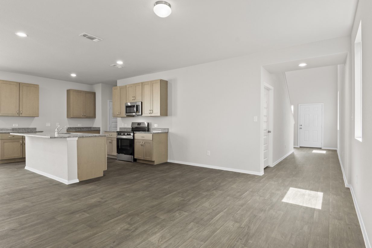 Interior image of kitchen with tan cabinets and granite countertops and living room with vinyl flooring.