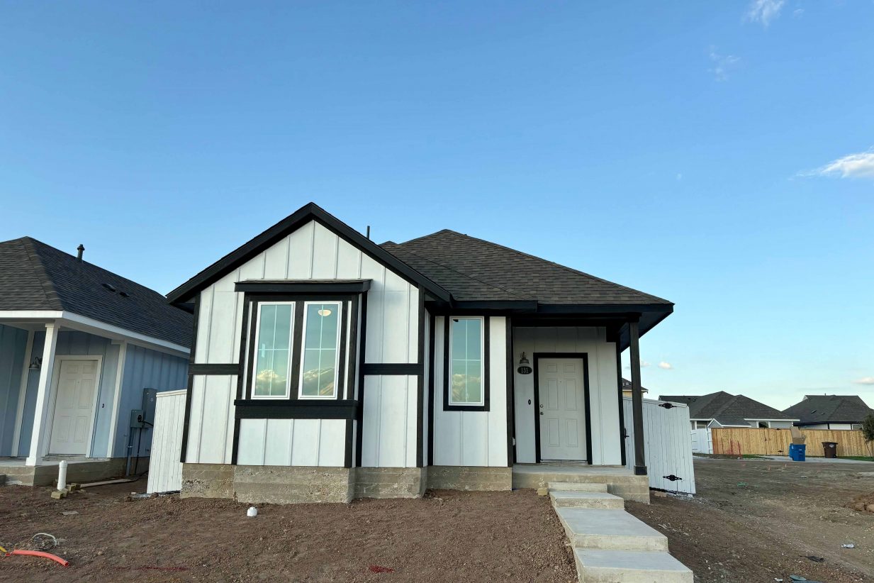Image of front of a white farmhouse-style single story home with black trimming on a dirt lot and blue sky in the background.