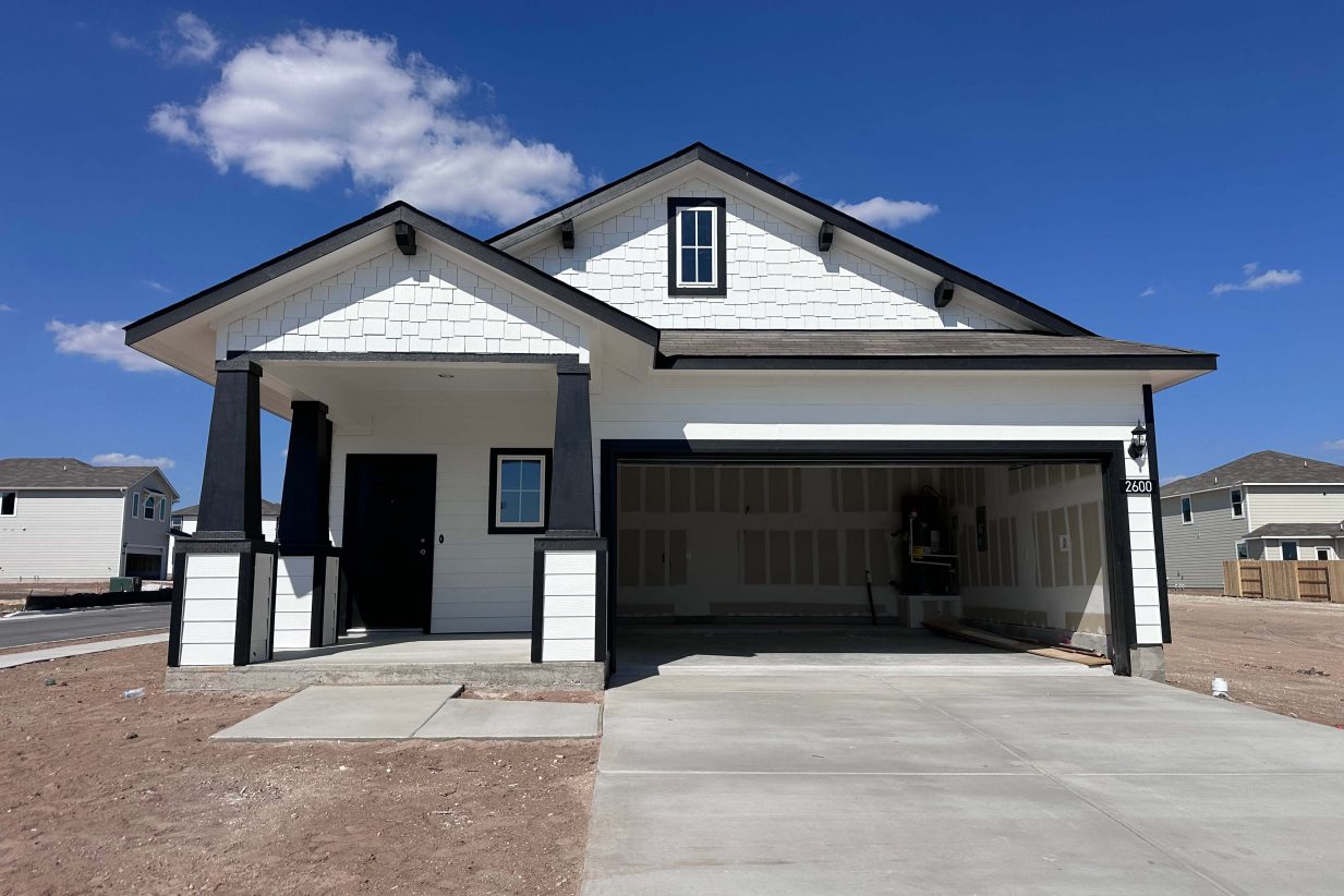 Image of front of white single-story house with black trimming, a driveway, dirt landscape, and a blue sky in the background.