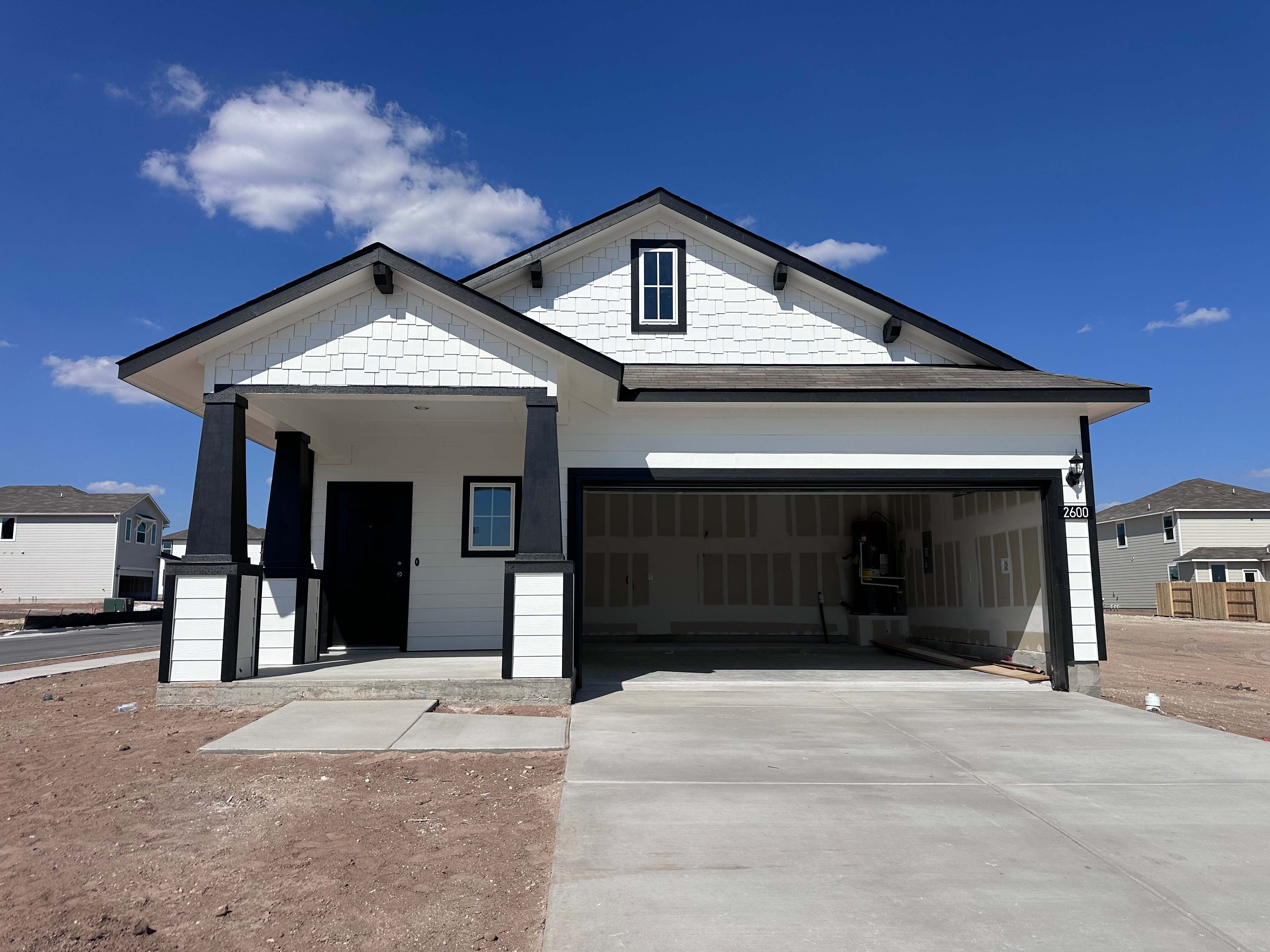 Image of front of white single-story house with black trimming, a driveway, dirt landscape, and a blue sky in the background.