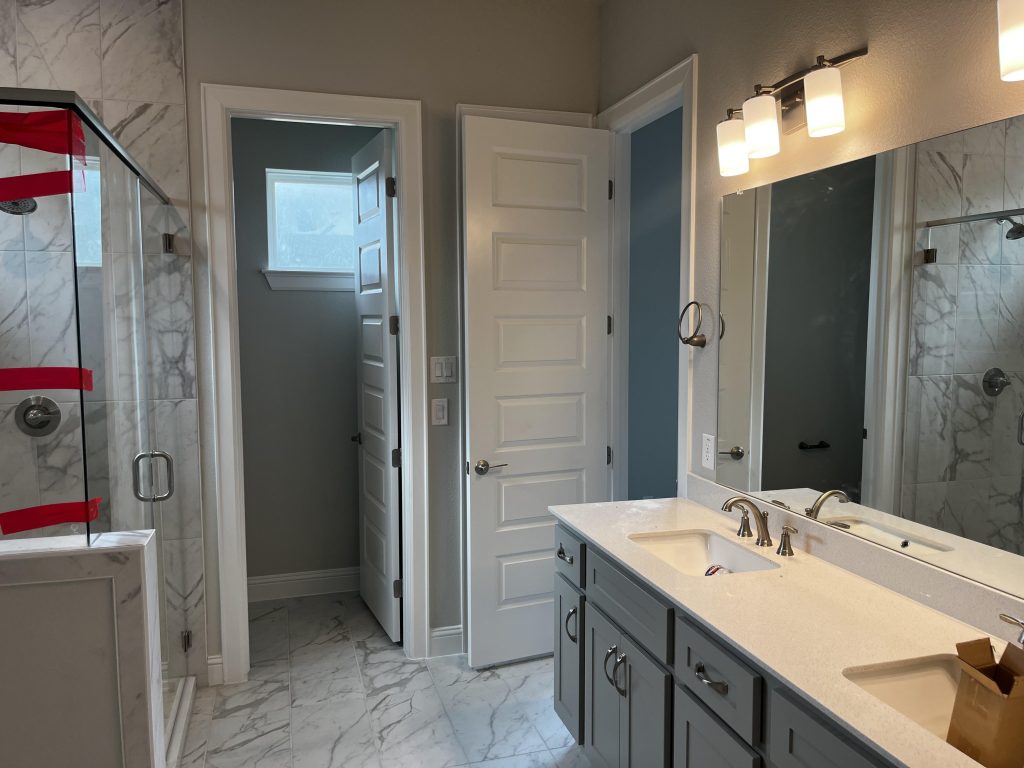 An image of bathroom with a marbled tiled floor, a glass shower with marbled tiled wall, and dual vanity with white countertop and grey cabinets.