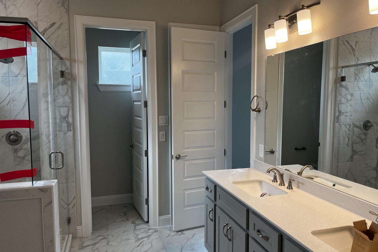 An image of bathroom with a marbled tiled floor, a glass shower with marbled tiled wall, and dual vanity with white countertop and grey cabinets.