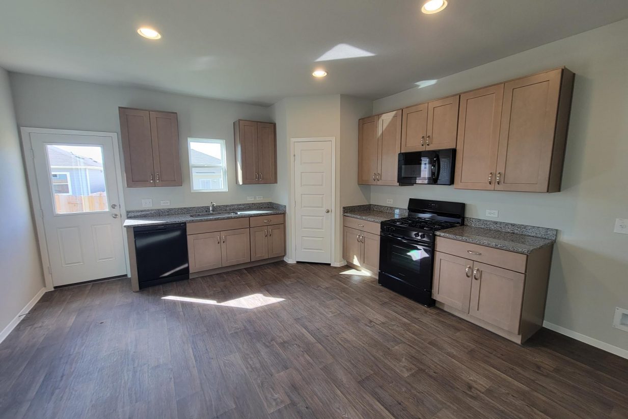 Image of home kitchen with wood-look floor, light brown cabinets, window over sink and in rear door, white walls, and black appliances