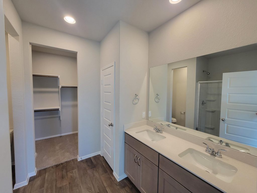 Image of home primary bathroom with dual vanity with white countertop and light brown cabinets, mirrow, white walls, wood-look floor, and doorway to walk-in closet