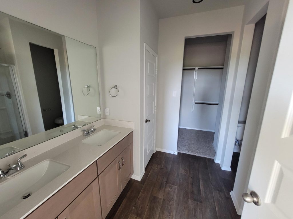 Image of home bathroom with double vanity with light brown cabinets and white countertops, mirror, and doorway to walk-in closet