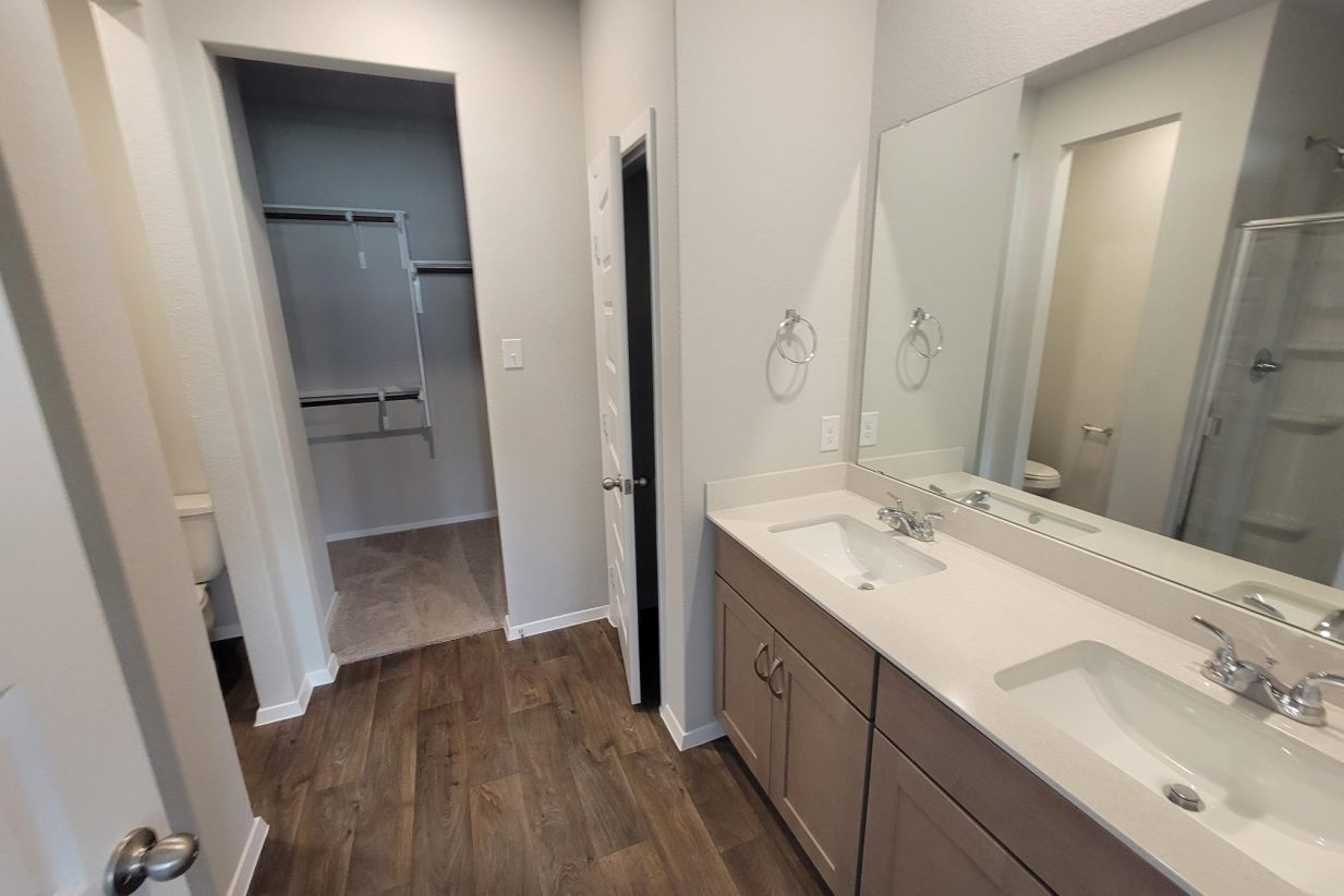 Image of home bathroom with dual vanity with white countertop and light brown cabinet, mirror, wood-look floor, and doorway to walk-in closet