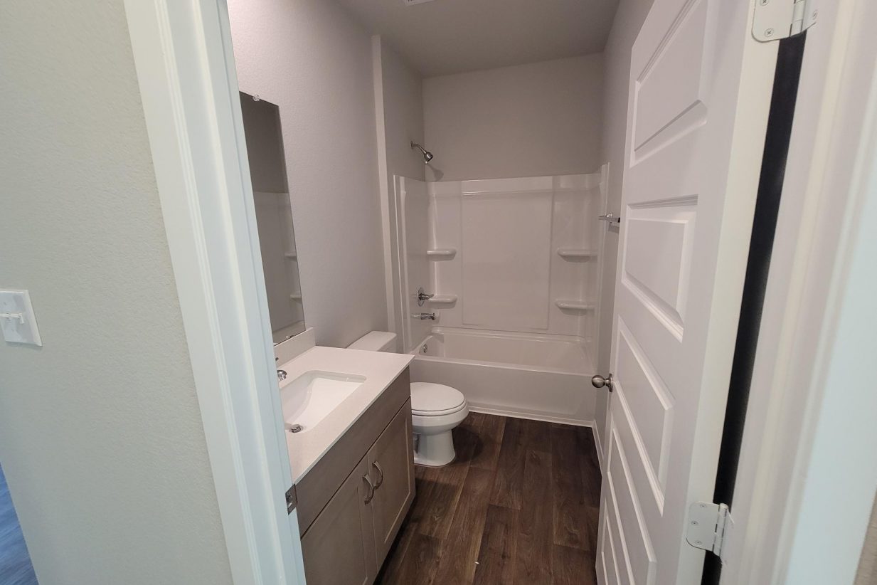 Image of home bathroom with vanity with white countertop and light brown cabinet, toilet, tub, wood-look floor, mirror, and white walls