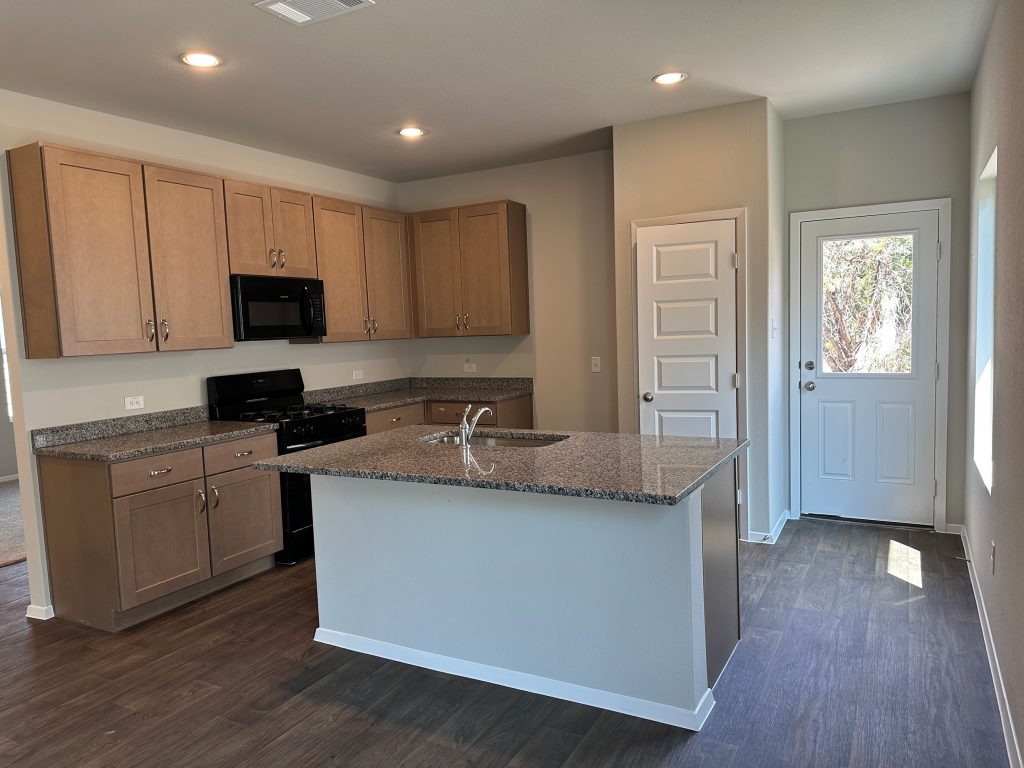Image of home kitchen with light brown cabinets, center island with sink, pantry door, and rear door with window. Light wood-look flooring and light walls