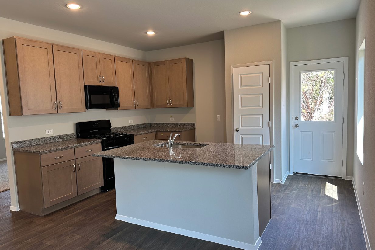 Image of home kitchen with light brown cabinets, center island with sink, pantry door, and rear door with window. Light wood-look flooring and light walls
