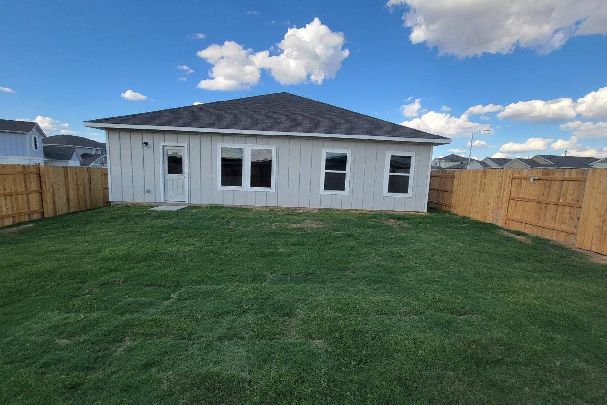 Image of one-story home exterior with green grass, wood fence, and sky