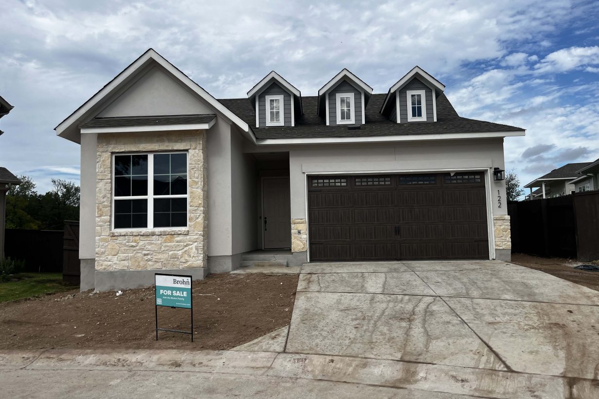 Front exterior of gray and brick accented single-story house with a brown two-car garage and dirt landscape.