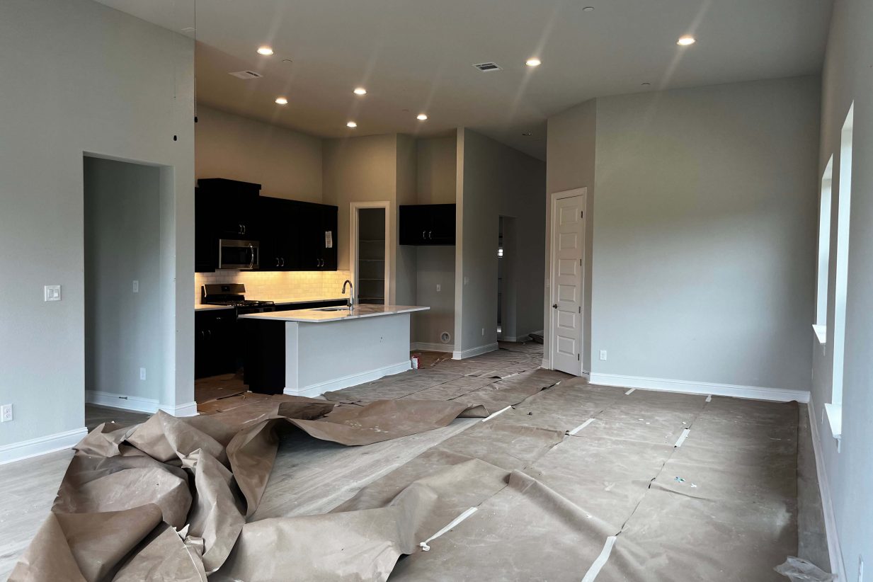 A kitchen and living room with white marbled countertops, brown cabinets, grey painted walls, and white framed base boards.