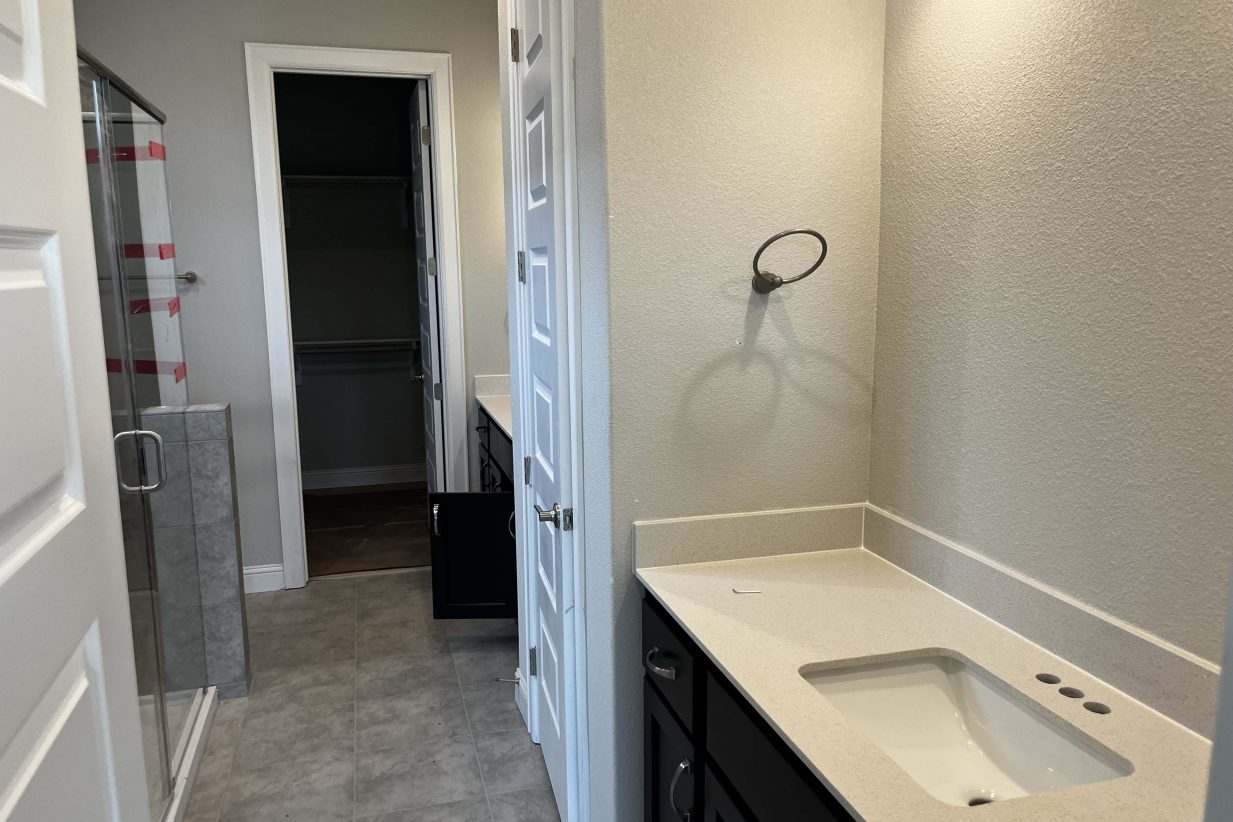 A bathroom with gray floor tiles, a glass shower, a vanity with white countertop and brown cabinets and gray painted walls.