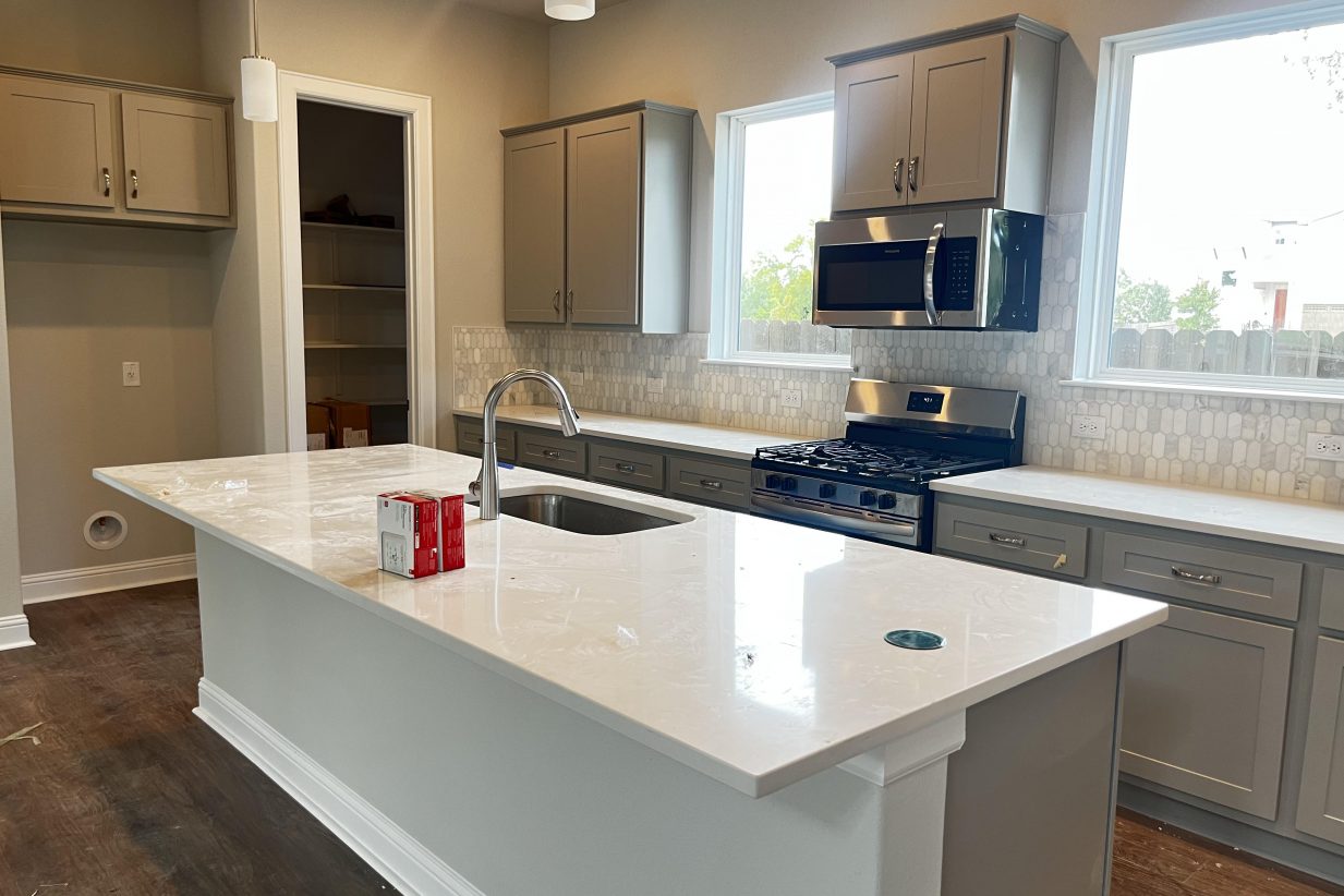 An image of a kitchen with brown vinyl flooring, white and grey backsplash tiling, grey cabinets, and white countertops, a central island with white countertop, and steel appliances.